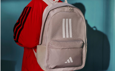 A gray Adidas backpack with three white stripes is shown against a teal background, with a red jacket or clothing item visible in the foreground.