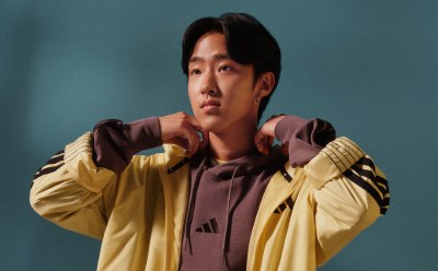 Mladý Ázijčan v žltej bunde a fialovej košeli stojí na modrom pozadí, jeho vyjadrenie je premyslené a sústredené.