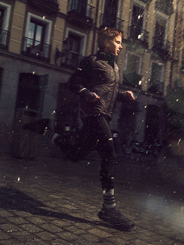 A person in a dark jacket stands in a snowy urban setting, with buildings and streetlights visible in the background.