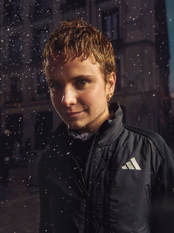 A young man with blond, tousled hair stands in the foreground, wearing a black Adidas jacket, against a dark background with falling snow.
