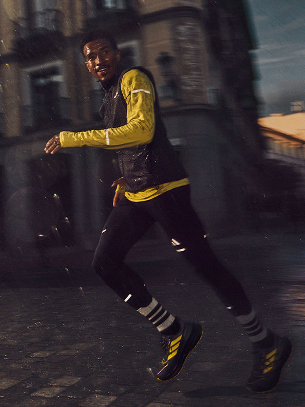 A person in a yellow jacket and dark pants is running through a dimly lit urban environment, with blurred buildings and structures in the background.