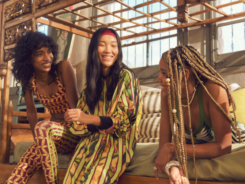 Three women with long, dark hair and vibrant, patterned clothing sit together in a cozy, rustic-looking indoor space with wooden beams and large windows.