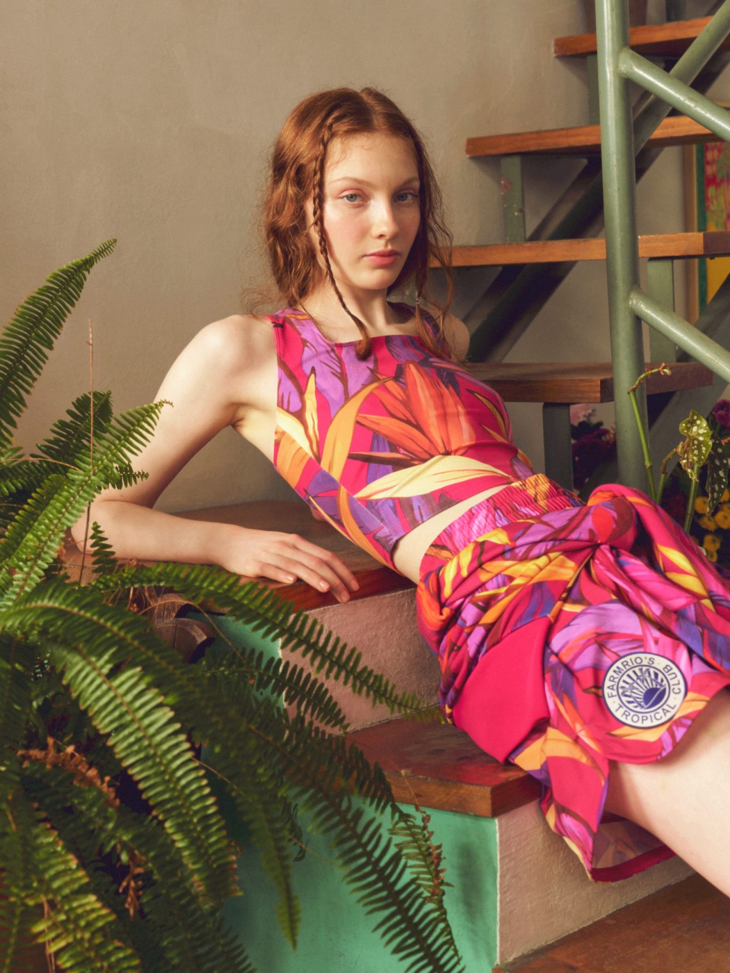 A young woman with curly hair reclines on a chair surrounded by lush greenery, wearing a vibrant, patterned dress.