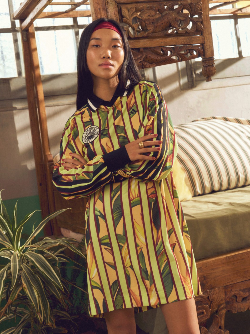 A young woman wearing a vibrant, patterned dress stands in a rustic, wooden-framed room with a potted plant in the background.