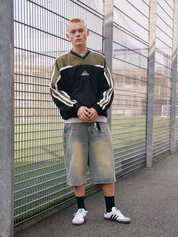 A man in an adidas long sleeve top, grey shorts, and white adidas Samba shoes casually poses against the fence of a tennis court.