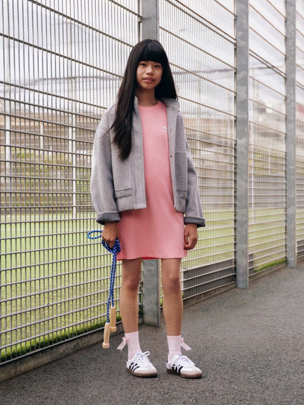 A girl with black hair wears a pink dress, beige jacket, and white Samba shoes while standing outside the fence of a tennis court.