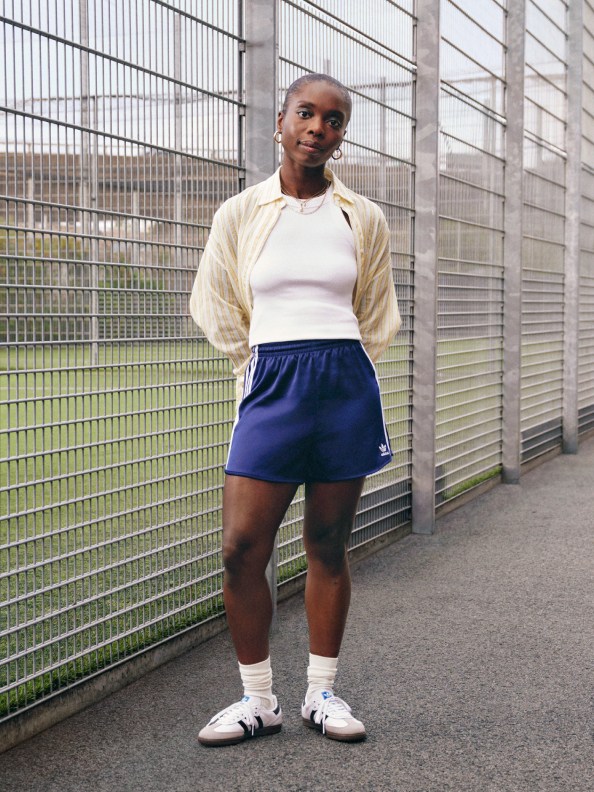 A woman in a blue adidas skirt, white tee, and yellow cardigan poses with her hands behind her back in front of a tennis court.