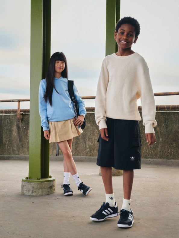 A girl and a boy are all smiles as they pose in adidas apparel and black SL 72 shoes on a concrete street.