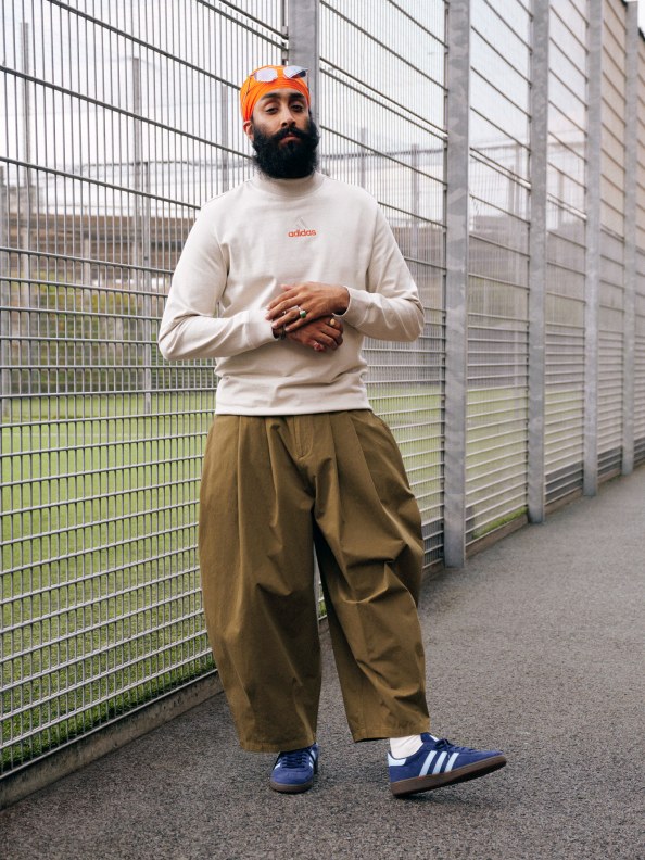 A woman in a black adidas tracksuit jacket, grey shorts, and white adidas Handball Spezial shoes poses in front of a tennis court fence. 