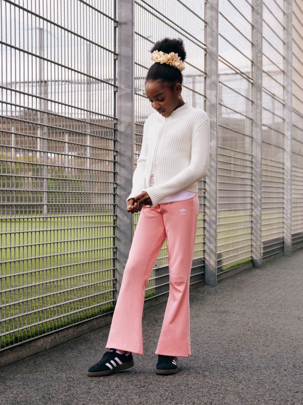 A girl in a white cardigan, pink pants, and black adidas Handball Spezial shoes poses and looks at the ground in front of a steel fence.