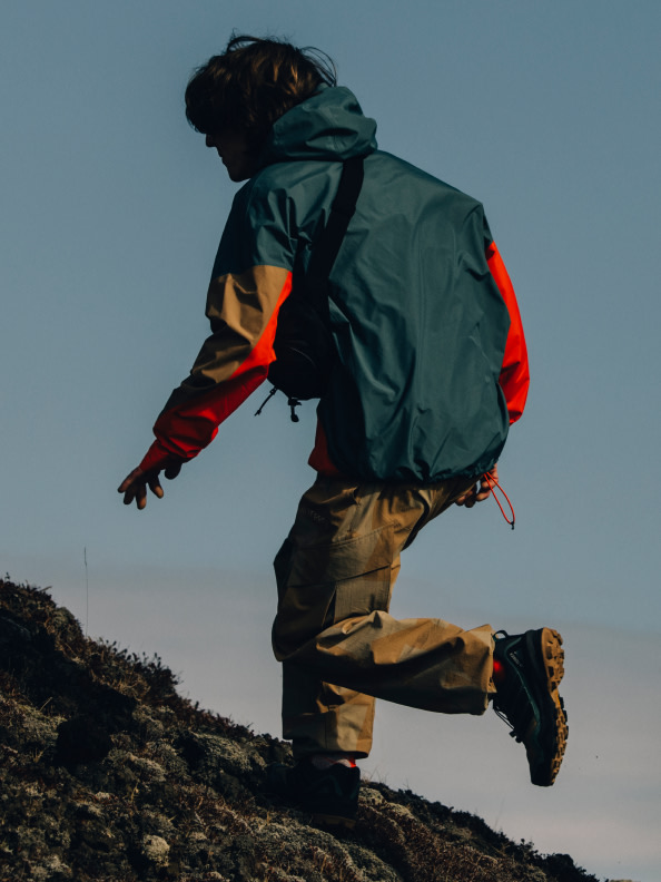 A person in a green and orange jacket and pants is hiking up a rocky hill against a clear sky backdrop.