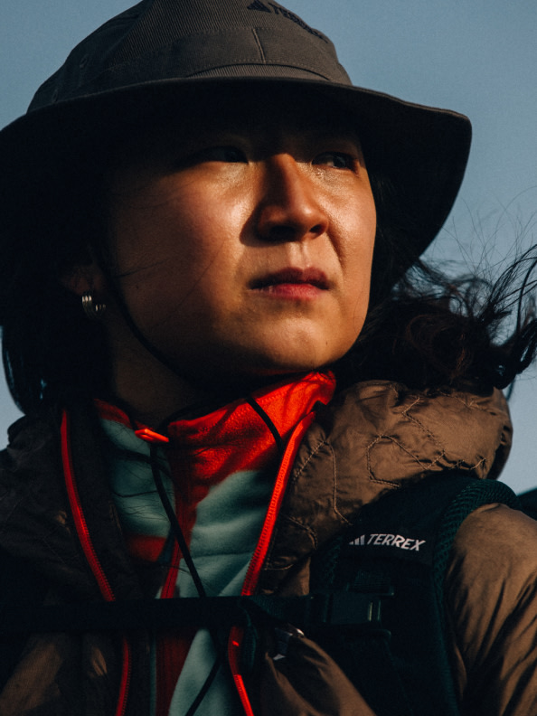 A person wearing a black hat and red jacket stands against a cloudy sky, their face partially obscured by the hat's brim.