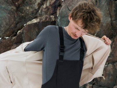 Ένα άτομο με μακριά, κυματιστά μαλλιά φορώντας γκρι πουλόβερ και μαύρες ολόσωμες φόρμες στέκεται μπροστά από ένα βραχώδες, ορεινό τοπίο.