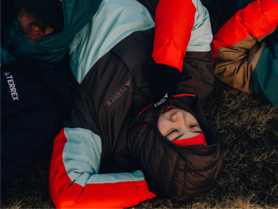 Η εικόνα απεικονίζει ένα άτομο να ξαπλώνει στο έδαφος, τυλιγμένο με σκούρα ρούχα και περιτριγυρισμένο από πολύχρωμα υφάσματα, δημιουργώντας μια εντυπωσιακή οπτική αντίθεση.