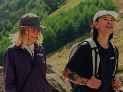 Dwie osoby, kobieta i mężczyzna, idą razem na szlaku otoczonym bujną, zieloną roślinnością i górskim krajobrazem w tle.