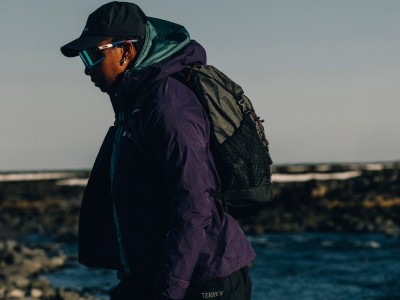 Une personne portant une veste à capuche sombre se tient dans un cadre extérieur pittoresque, avec une étendue d'eau et un paysage rocheux visible en arrière-plan.