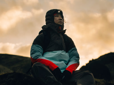 Une personne portant une veste à capuche noire et des vêtements colorés se tient sur un fond de ciel nuageux et doré et de terrain montagneux accidenté.