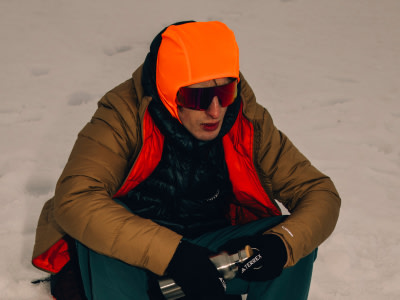 Une personne portant un chapeau orange vif, des lunettes de soleil et une veste de bronzage se tient dans un cadre extérieur enneigé.