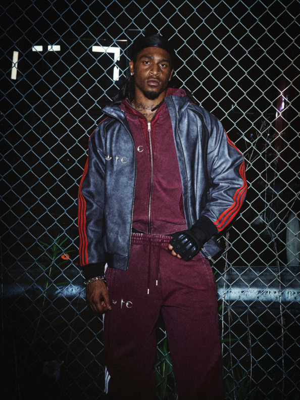 A man wearing a burgundy tracksuit and a navy blue leather jacket stands in front of a chain-link fence in a dimly lit outdoor setting.