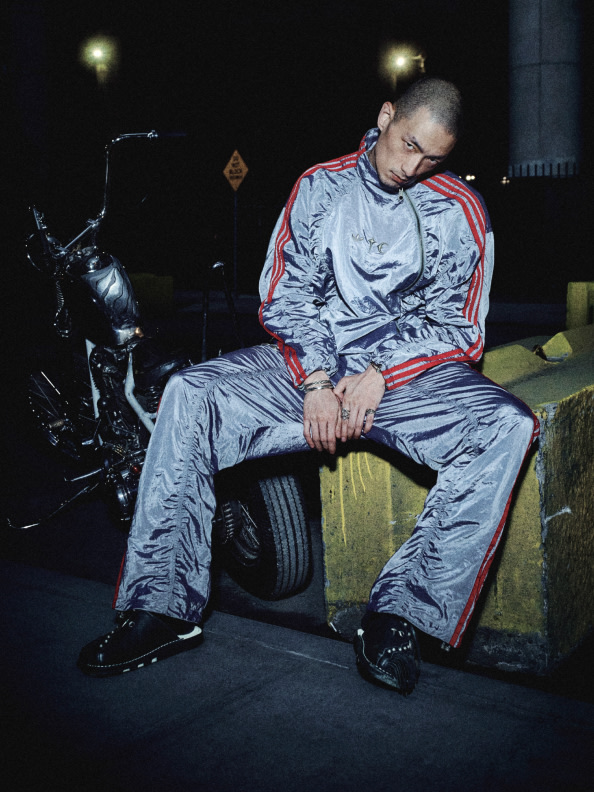 A person wearing a silver and red tracksuit is sitting on a tyre in a dimly lit outdoor setting, with a motorcycle visible in the background.