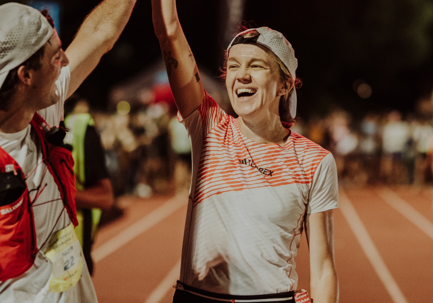 The image shows two people, one in a red shirt and the other in a white and red striped shirt, celebrating and cheering on a sports field with a crowd in the background.