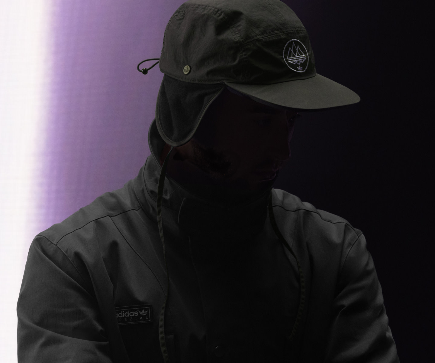 A person wearing a black hooded jacket and cap stands in a dimly lit, purple-tinted environment.