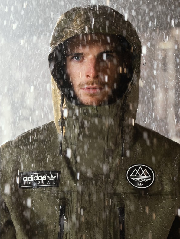 adidasとmountainのロゴをあしらったフード付きジャケットを着た人物が、雨の中に立つ。フードに顔の一部が隠れている。