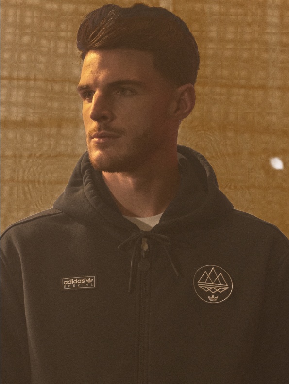 無地の背景に、adidasのロゴと山のシンボルを掲げた黒いフード付きジャケットを着た若者が立っている。