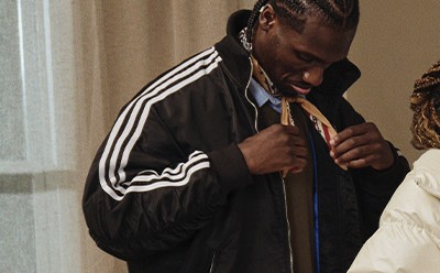 A person wearing a black jacket with white stripes stands in a room with a curtain visible in the background.