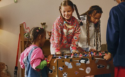 A group of young people, including two girls with braided hair, are gathered around a table playing a game or engaging in some activity together in what appears to be a cozy indoor setting.