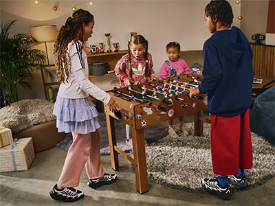 A group of children, two girls and two boys, are gathered around a foosball table, engaged in a lively game, with a cozy and decorated living room setting in the background.