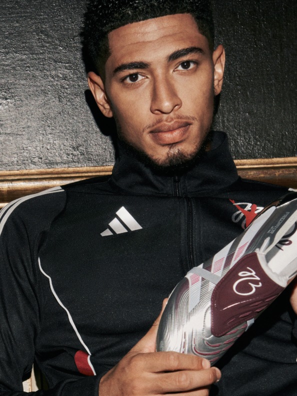 Jude Bellingham wearing a black athletic jacket and holding a soccer ball stands in front of a dark background.