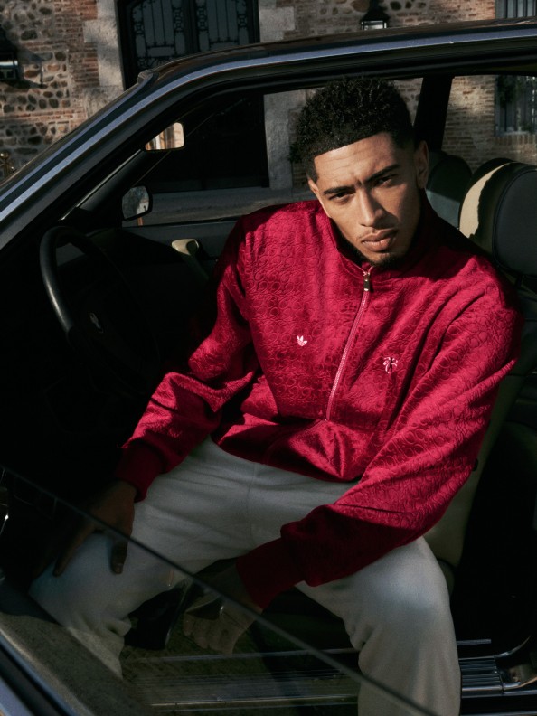Jude Bellingham wearing a red jacket sits in the backseat of a car, with a brick building visible in the background.