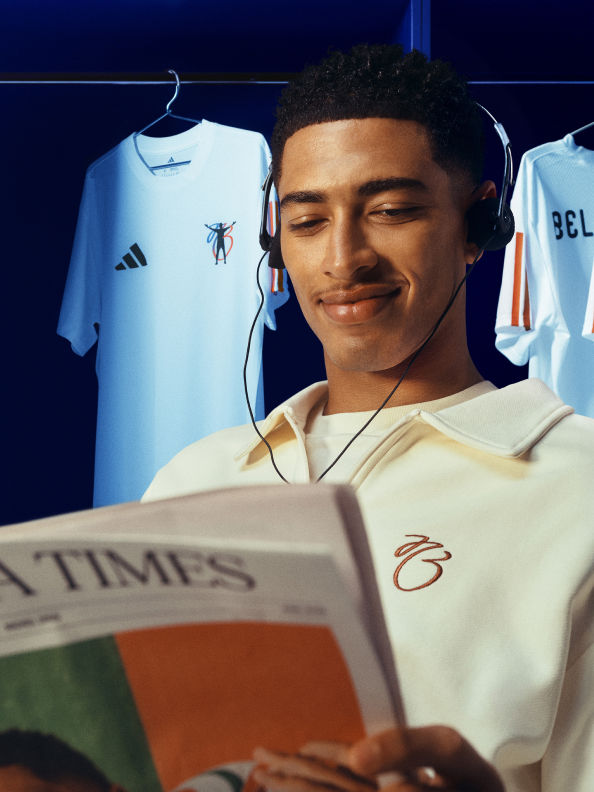 A young man with a warm smile is reading a newspaper while surrounded by sports jerseys and equipment in the background.