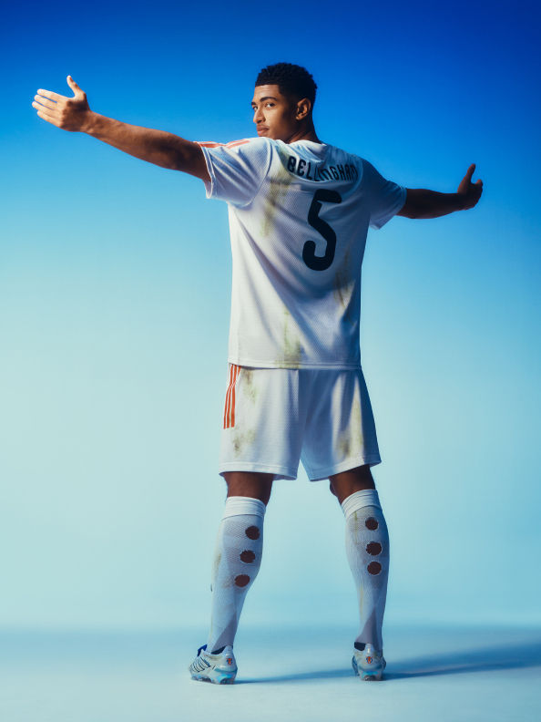 A young soccer player in a white uniform stands with arms outstretched against a clear blue sky.
