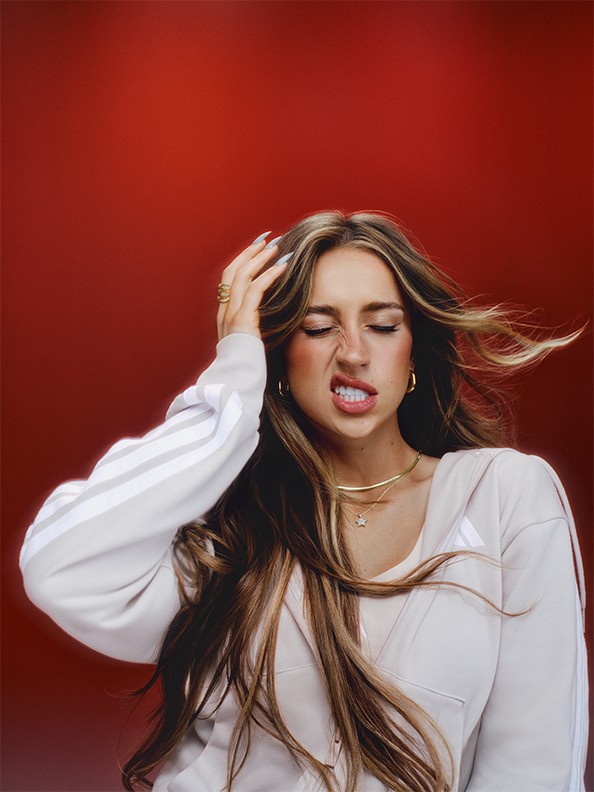 A young woman with long, flowing brown hair appears to be in a state of distress, her eyes closed and her hands grasping her head, against a vibrant red background.