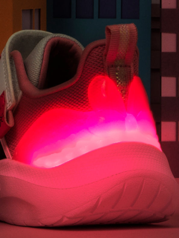 선반 및 기타 물체가 보이는 희미한 조명 배경과 대비된 앞발에 반짝이는 아웃솔과 텍스처드 갑피가 있는 생기 넘치는 네온 핑크 색상의 운동화가 있습니다.