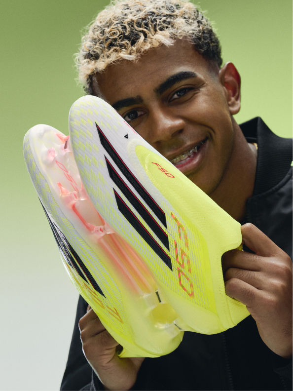 A smiling young man with curly hair holds a bright yellow and pink sports shoe in front of his face against a green background.