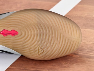 The image shows a close-up view of a wooden kitchen utensil, likely a potato masher or ricer, with a distinctive wavy pattern on its surface and a red silicone accent. The utensil is resting on a wooden surface, possibly a kitchen counter or table.