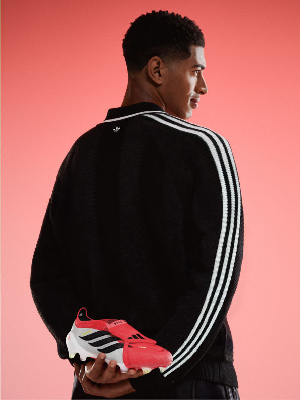 A young man wearing a black jacket with white stripes stands against a vibrant red background, holding a pair of red and white athletic shoes.