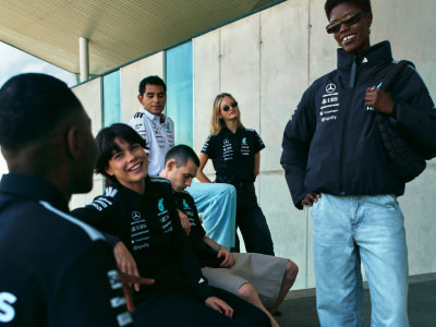 A group of young people, some smiling and others with serious expressions, stand together in an indoor setting with a tiled wall in the background.