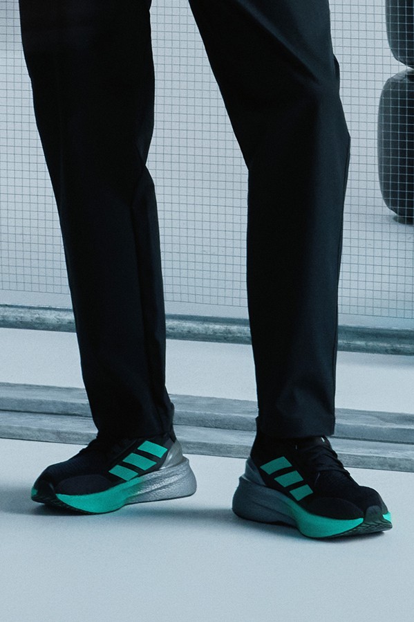 The image shows a person's lower body wearing black pants and a pair of black and teal athletic shoes standing in front of a white tiled wall.