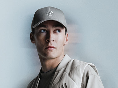 Mladý muž v sivej bejzbalovej šiltovke s logom na nej sa pozerá priamo na fotoaparát s vážnym výrazom na čisto modrom pozadí.