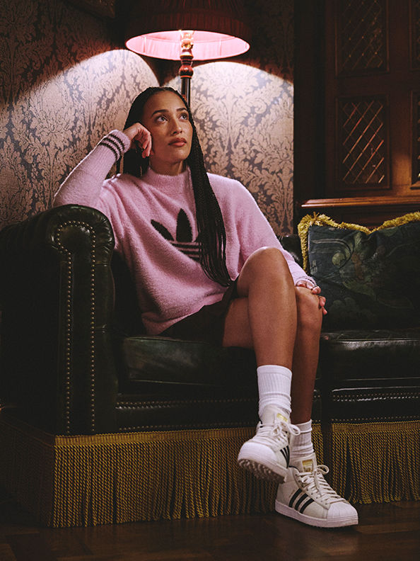 Una mujer joven con una sudadera rosa y calcetines blancos sentada en una silla decorada y acolchada, sosteniendo un paraguas rosa sobre su cabeza en un decorado interior oscuro y adornado.