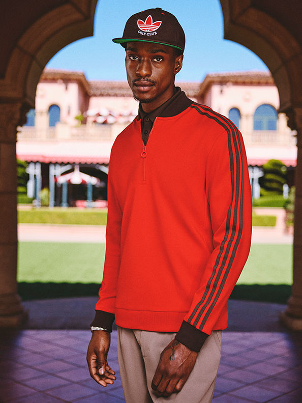 Un joven con una chaqueta roja de adidas y una gorra negra de béisbol se coloca de pie frente a un colorido edificio decorado con entradas arqueadas.
