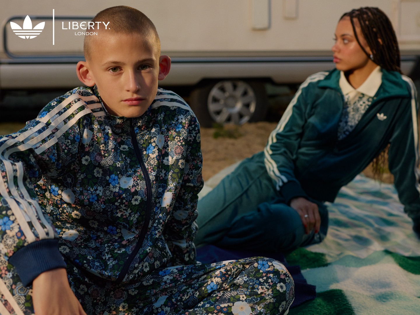 A boy and a girl sitting in front of a caravan. He is wearing a blue and white adidas floral outfit, and she is wearing a green adidas tracksuit. 
