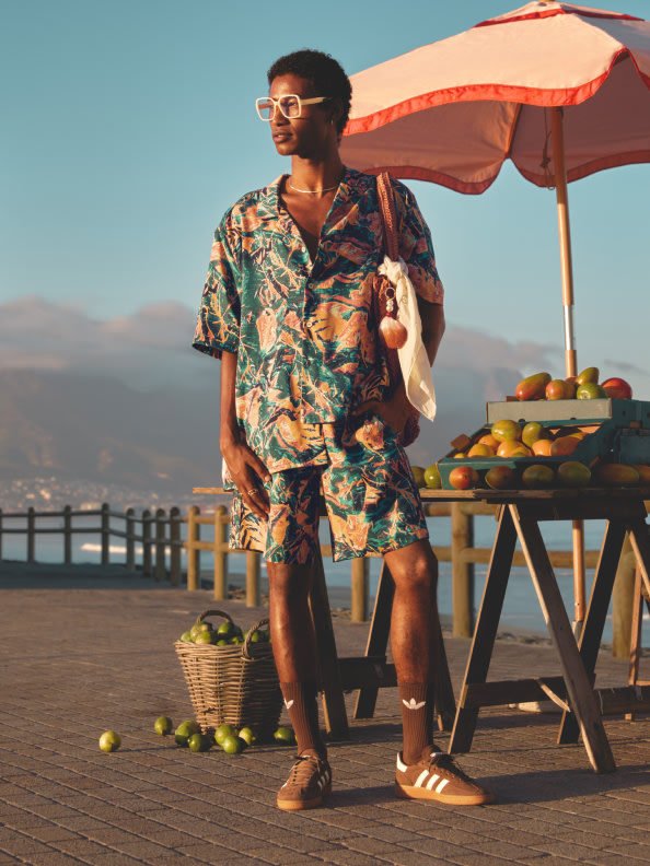 A person wearing a colorful, patterned shirt and shorts stands in front of a fruit stand on a wooden boardwalk, with a beach and mountains visible in the background.