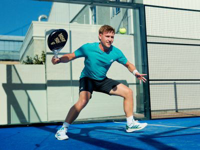 A man in a teal shirt and shorts is playing a game of padle tennis on a blue court.