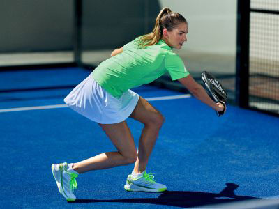 A young woman in a bright green top and white skirt is running on a blue padel court.
