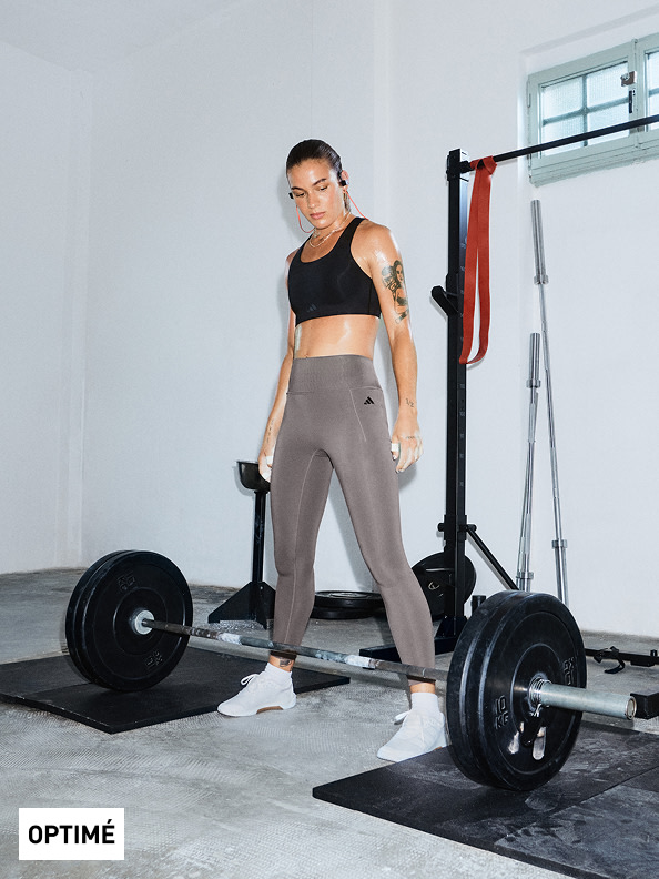 Una persona con un sujetador deportivo negro y mallas grises está de pie en el gimnasio, con equipamiento de ejercicios como una barra y una barra de pesas visibles en el fondo.